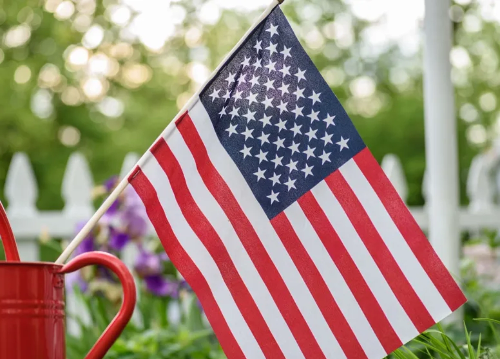 cr=w_776,h_583 (3)_11zon A small American flag is placed in a red container in a garden with a white fence and foliage in the background.